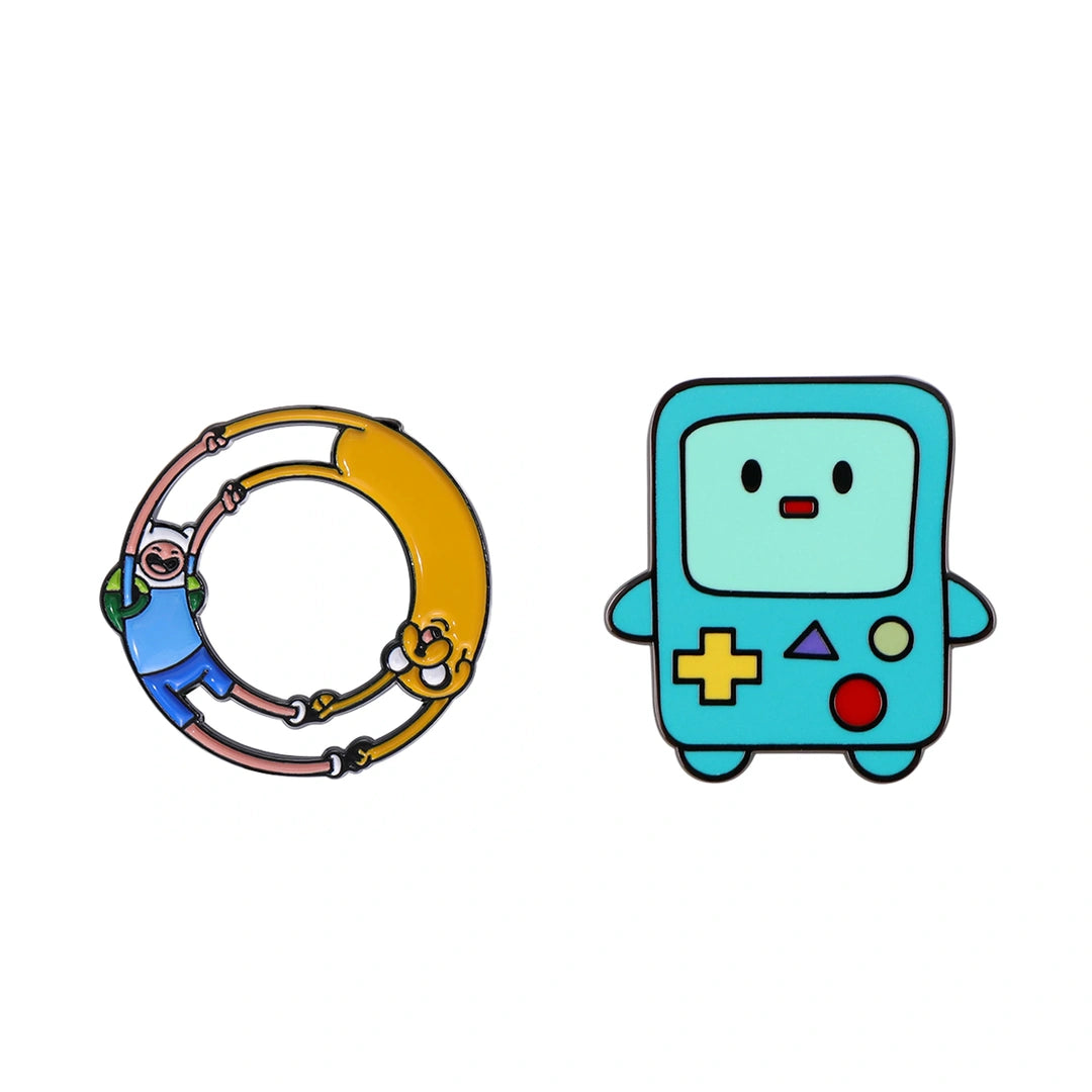 Two cartoon enamel pins on a plain white background: a yellow ring pin featuring Adventure Time characters Finn and Jake, and a light blue game-controller-shaped pin resembling BMO.