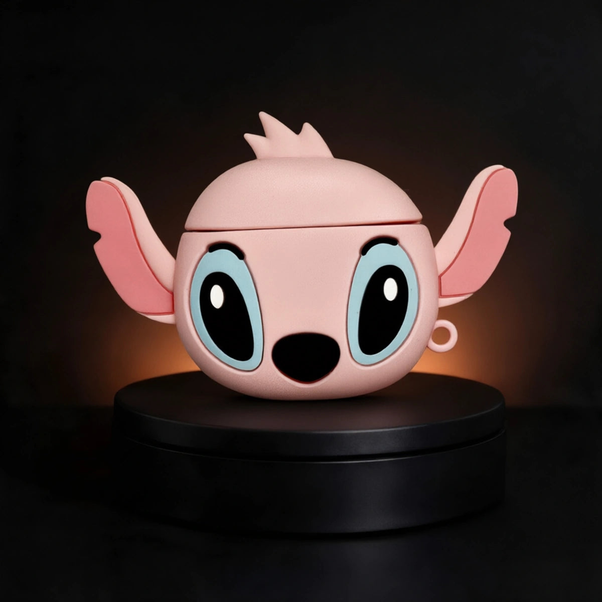 A pink character-shaped as angel from stitch AirPods case designed to look like a cartoon creature with large blue eyes, pink ears, and a black nose, displayed on a black circular pedestal against a dark background.