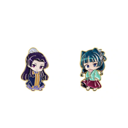 Two small chibi-style enamel pins on a plain white background. The left pin shows a dark-haired character in a navy and gold outfit, and the right pin shows a teal-haired character in a light green and burgundy outfit, both outlined in gold.