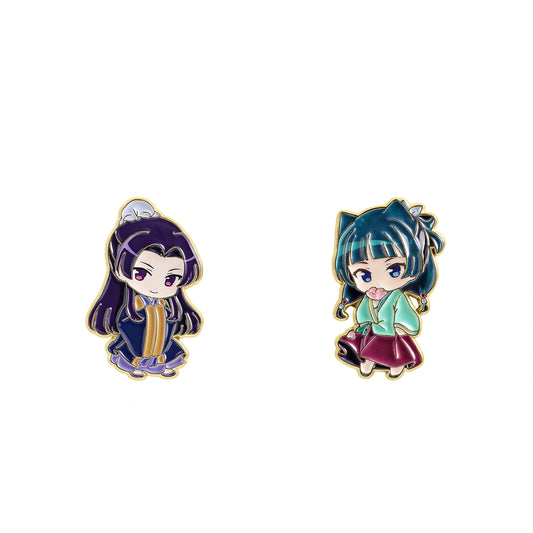 Two small chibi-style enamel pins on a plain white background. The left pin shows a dark-haired character in a navy and gold outfit, and the right pin shows a teal-haired character in a light green and burgundy outfit, both outlined in gold.