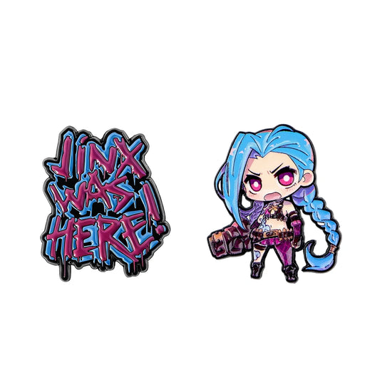 Two enamel pins on a plain white background: one features stylized graffiti text reading 'JINX WAS HERE!' in pink and blue, and the other shows a chibi anime-style character with bright blue hair, pink eyes, and purple clothing holding a camera.