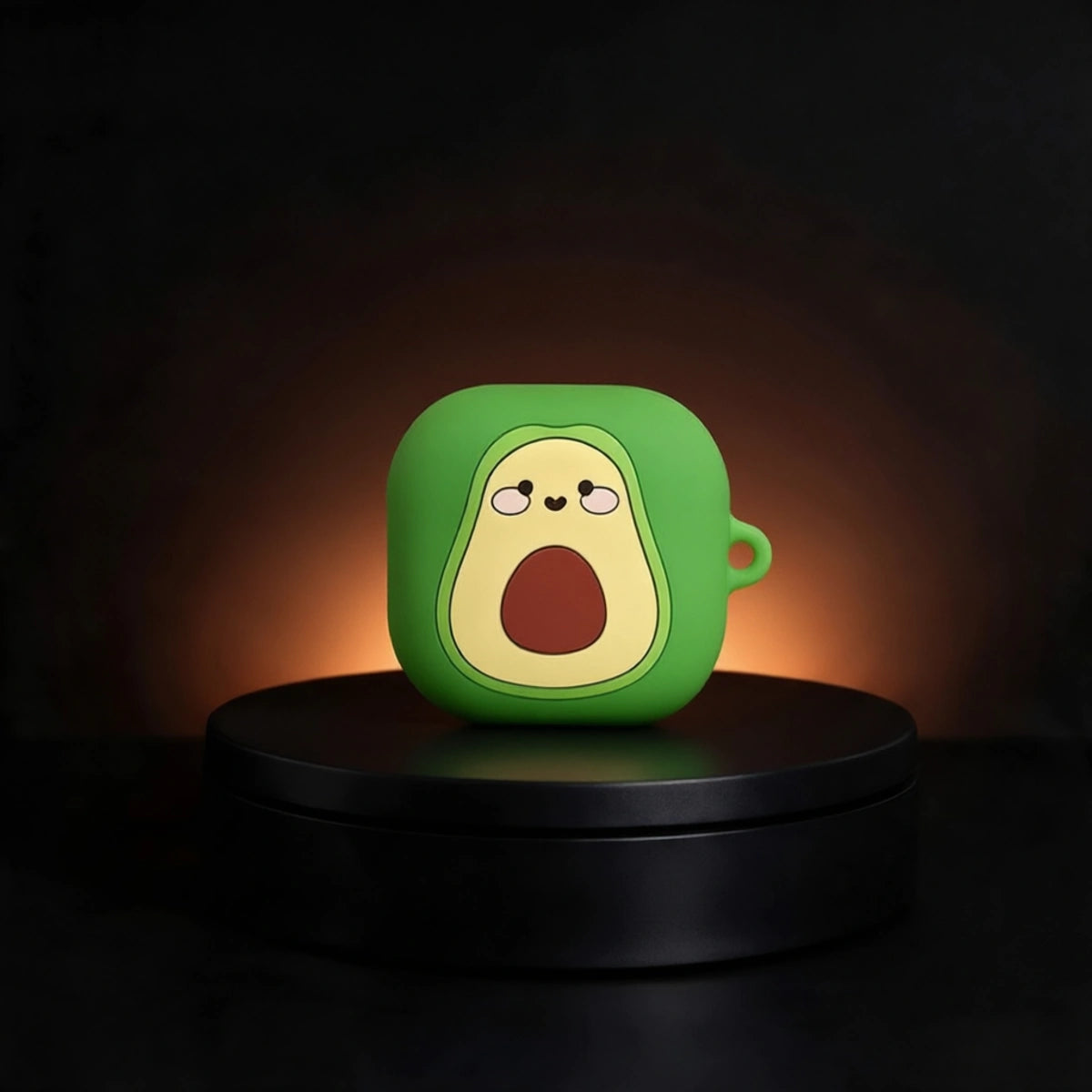 A green avocado galaxy buds case with a cartoon avocado face, displayed on a black circular pedestal against a dark background with warm lighting.