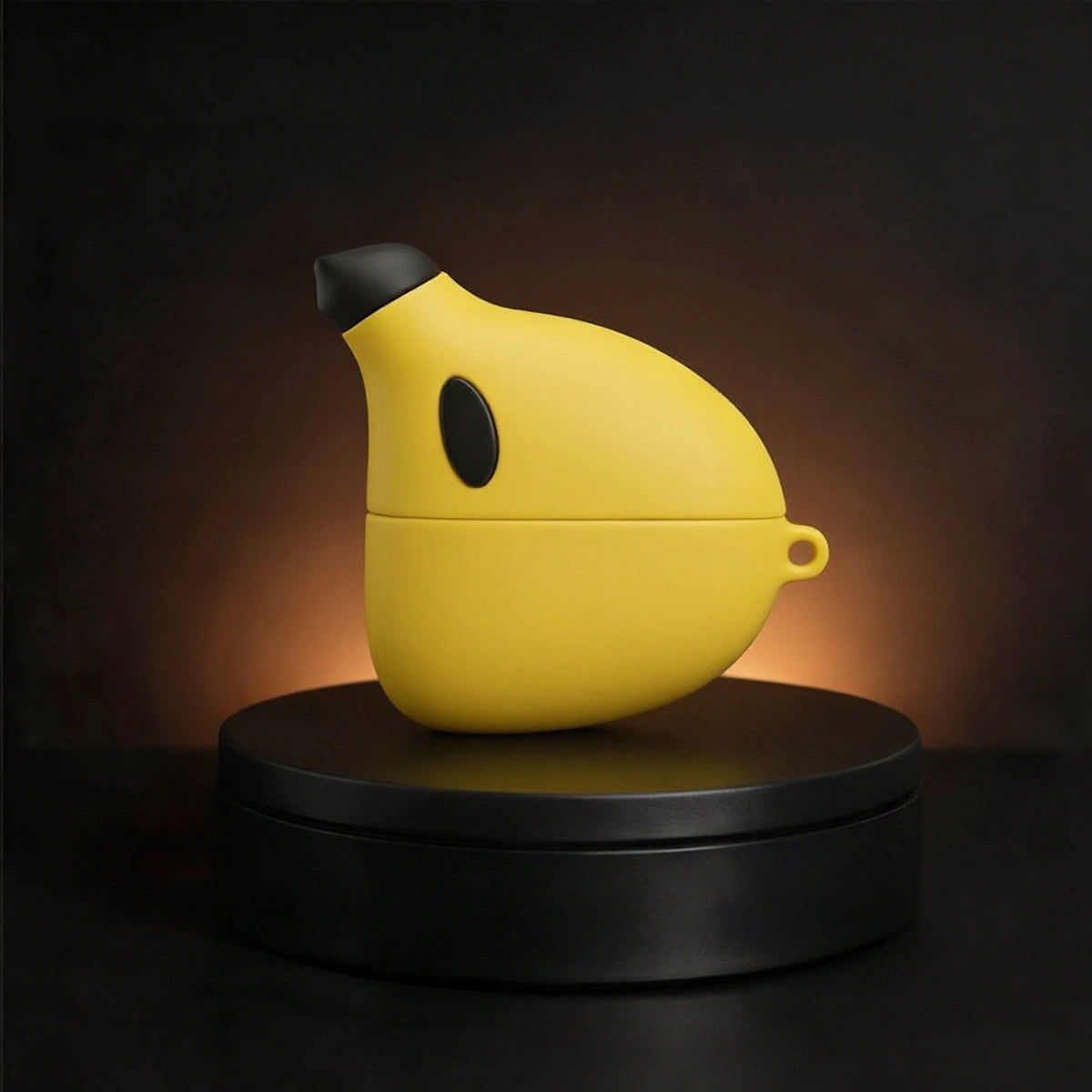 A yellow banana-shaped wireless earbud charging case with a black nose and side detail, displayed on a black circular pedestal against a dark background with warm light.
