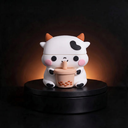 A cute cartoon cow airpods case with orange horns and a black heart-shaped patch on its forehead, holding a cup of bubble tea. The figurine is displayed on a black circular pedestal against a dark background with a warm glow behind it.