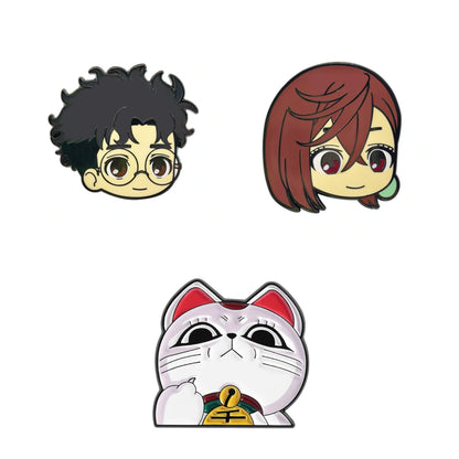 Three cartoon-style enamel pins on a plain white background: a black-haired round-eyed character at top left, a reddish-brown-haired character at top right, and a white maneki-neko cat with a gold bell at bottom center.