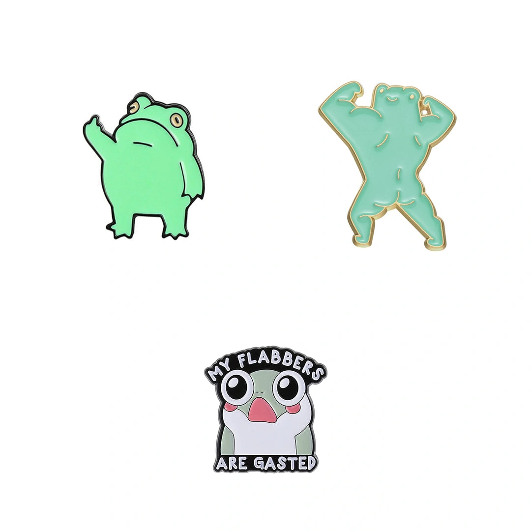 Three frog-themed enamel pins on a plain white background: a light green frog raising one paw, a mint green flexing frog outlined in gold, and a white and gray frog with pink cheeks and the text 'MY FLABBERS ARE GASTED' in black.