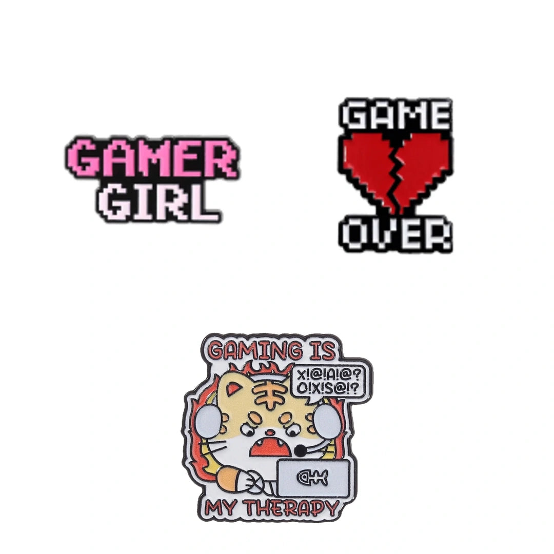 Three gaming-themed enamel pins on a plain white background: a pink and white pixel-style 'GAMER GIRL' pin, a red broken-heart pixel pin with black text reading 'GAME OVER,' and a multicolor cartoon pin showing a tiger character holding a laptop with the text 'GAMING IS MY THERAPY' and a speech bubble.