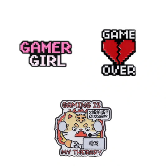 Three gaming-themed enamel pins on a plain white background: a pink and white pixel-style 'GAMER GIRL' pin, a red broken-heart pixel pin with black text reading 'GAME OVER,' and a multicolor cartoon pin showing a tiger character holding a laptop with the text 'GAMING IS MY THERAPY' and a speech bubble.