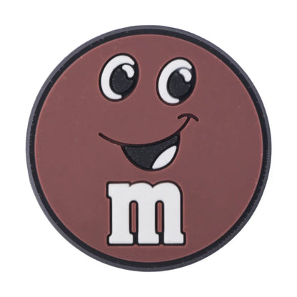 Brown M&M croc charm with a smiley face on a white background
