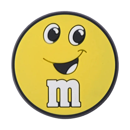 Round yellow M&M croc charm with a smiling face and the letter 'm' on a white background