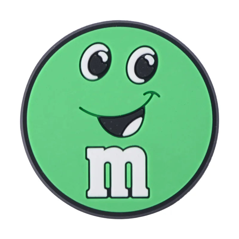 Green M&M croc charm with a smile on a white background