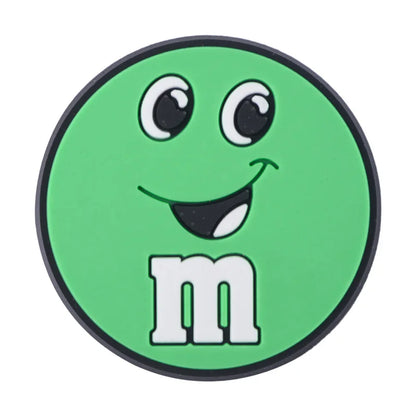 Green M&M croc charm with a smile on a white background