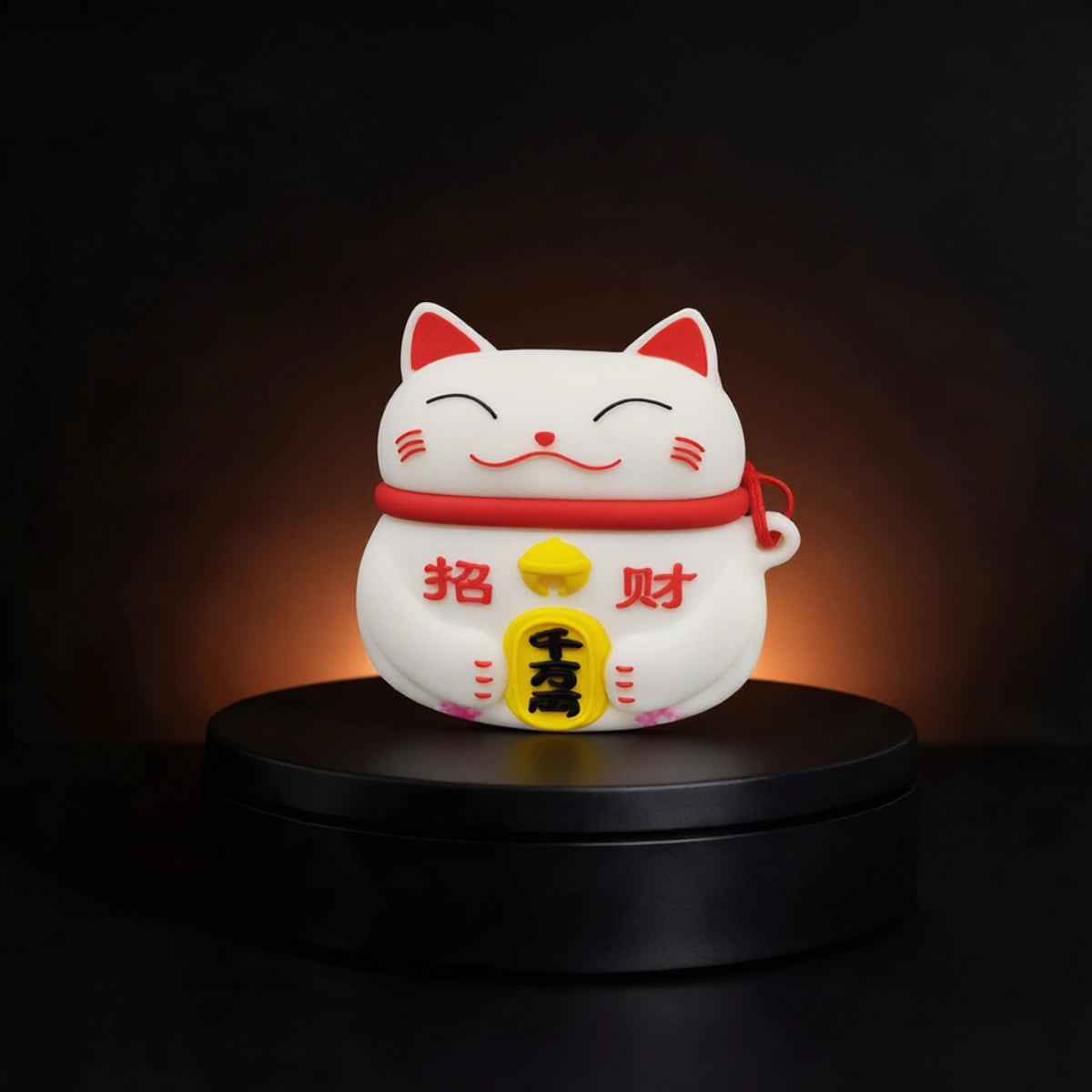 A white maneki-neko lucky cat airpods and buds case from inkbee store with red and yellow details, illuminated from within, displayed on a black circular pedestal against a dark background.