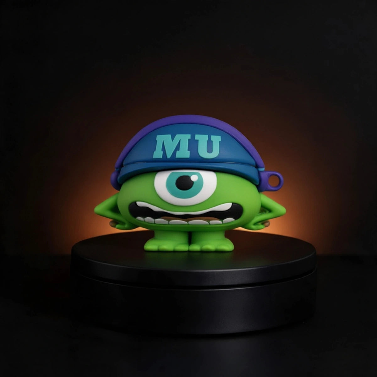 A green one-eyed cartoon character mike wazowski from monsters inc airpods and buds case figurine wearing a blue cap with 'MU' in light blue, standing on a black circular base against a dark background with a warm glow.