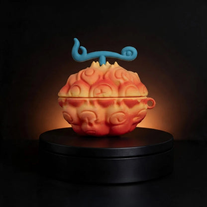 A small fruit-shaped decorative case for apple airpods and samsung galaxy buds with a blue swirl handle, shown on a black round pedestal against a dark background. The one piece flame flame devil fruit has a mottled orange, yellow, and red surface with embossed facial details.
