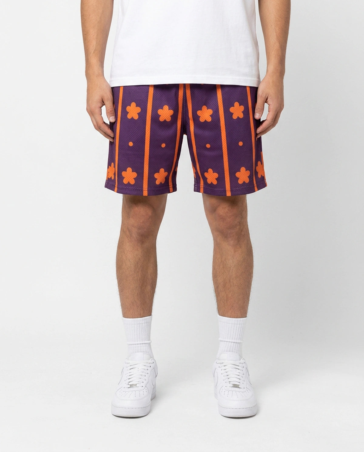 A person wearing patterned athletic shorts with a purple base, orange vertical stripes, and orange flower motifs, styled with a white t-shirt, white socks, and white sneakers against a light gray studio background.