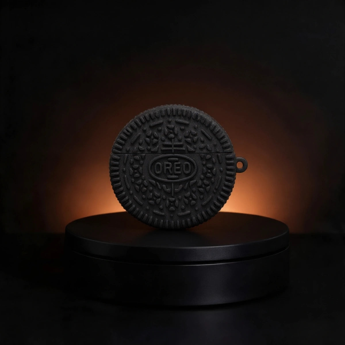 A black silicone case designed to look like an Oreo cookie, featuring the embossed OREO logo in the center, displayed on a black circular pedestal against a dark background with a warm orange glow.