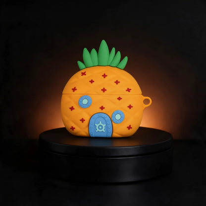 A pineapple-shaped wireless earbud case with a quilted texture, green leafy top, blue door, and red diamond details, displayed on a black circular pedestal against a dark background with warm light glow.