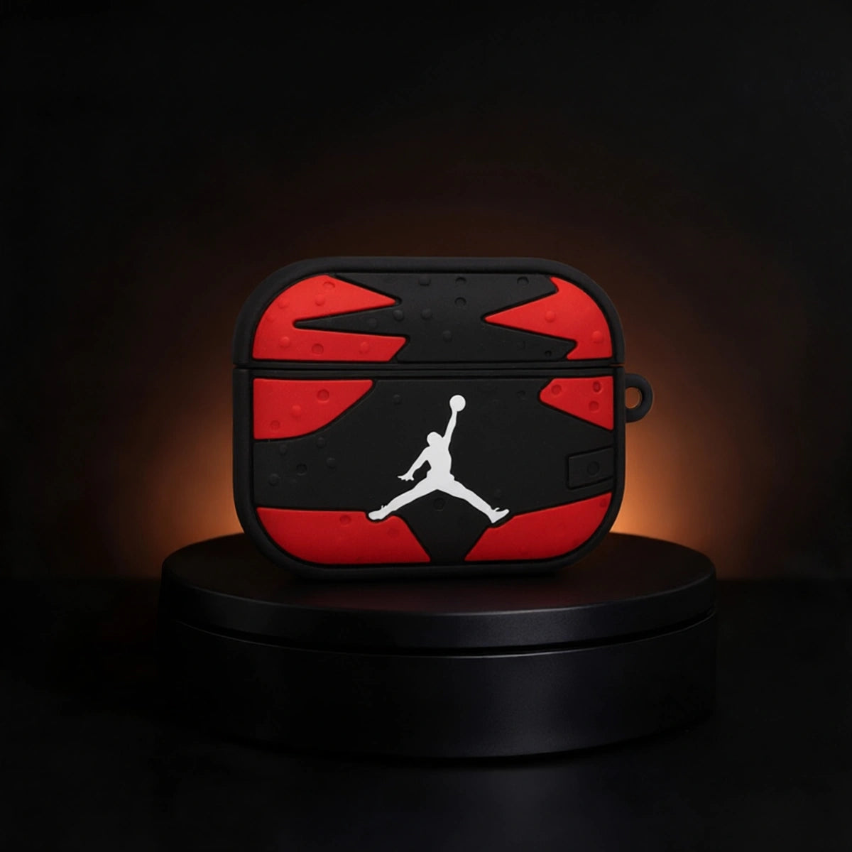 A black and red AirPods case with a white Jordan logo, displayed on a black circular pedestal against a dark background with warm lighting.