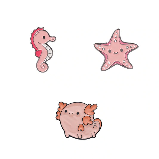 Three cute cartoon sea-themed enamel pins on a plain white background: a pink seahorse, a pink starfish, and a round pink axolotl.