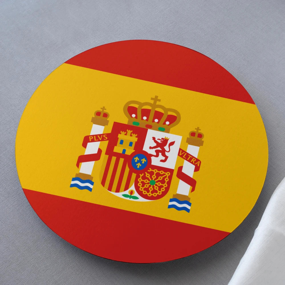 A round coaster featuring the Spanish flag design with its red and yellow horizontal bands and central coat of arms, photographed on a neutral gray surface.